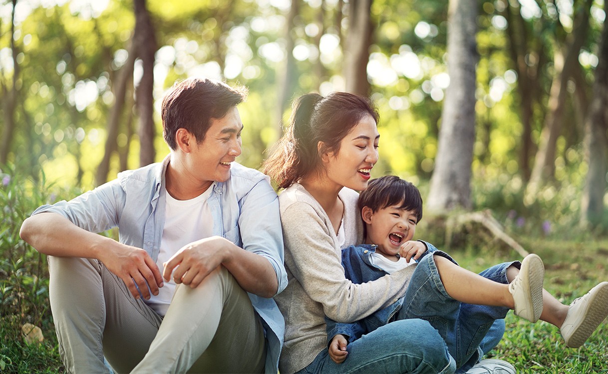 A happy family of three is enjoying a sunny day in the park. The mother sits on the grass with her legs crossed while holding her child close to her. Father sits to her side withan affectionate smile on his face. They both look at their child lovingly. In the background are tall trees providing shade from the sun's rays and lush green grass that stretches out for miles around them. This picture captures a moment of joy shared between this family as they spend quality time together outdoors in nature's beauty.