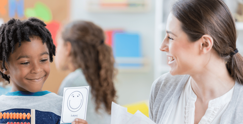 A young child smiles while holding up a card with a happy face drawing, as a therapist beside them smiles in encouragement. The setting is a bright, welcoming classroom with learning materials in the background. This image represents the My Emotions program at the SAAAC Autism Centre, which helps children identify, express, and regulate emotions using ABA-based strategies for emotional awareness and communication.