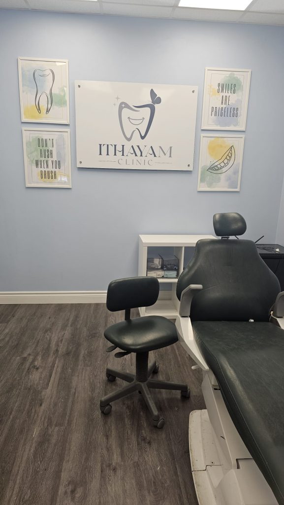 A clean and modern dental clinic room at Ithayam Clinic. The room features a black dental chair with an adjustable headrest, a matching black stool, and a white storage unit against a light blue wall. The clinic's logo, displaying a stylized tooth and the name 'Ithayam Clinic,' is prominently mounted on the wall. Surrounding the logo are framed motivational posters with watercolor designs, featuring messages like 'Don't rush when you brush' and 'Smiles are priceless.' The flooring is dark wood laminate, complementing the calming and professional ambiance.