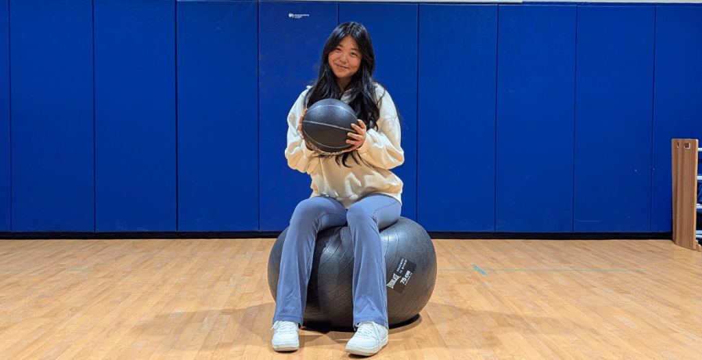 Ruby Bao, a volunteer with the SAAAC Autism Centre, sits on a large exercise ball in a gymnasium, holding a smaller medicine ball with a warm smile. She wears a cozy white sweater, light blue pants, and white sneakers. Behind her, blue padded gym walls provide a simple backdrop. Ruby supports autistic children through her volunteer work.