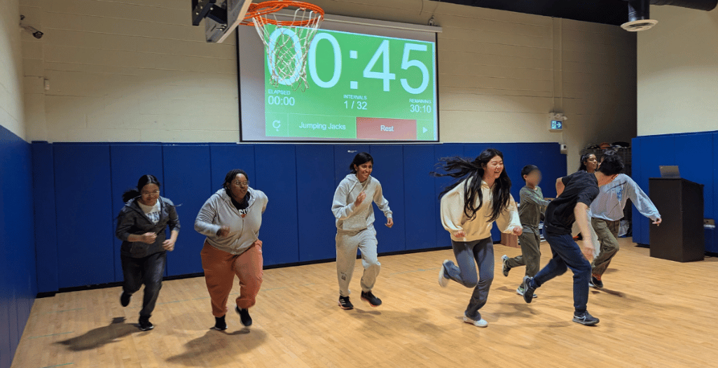 A group of volunteers and children, including Ruby Bao from the SAAAC Autism Centre, participate in a running exercise in a gymnasium. Ruby, wearing a cream-colored sweater and blue pants, is smiling as she runs alongside others. A large digital timer on the wall displays '00:45' with workout instructions. The participants, diverse in age and attire, are engaged in movement, bringing energy and enthusiasm to the activity.