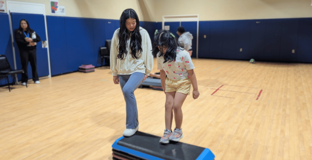 Ruby Bao, a volunteer with the SAAAC Autism Centre, supports a young girl during a step exercise in a gymnasium. Ruby, wearing a cream-colored sweater and blue pants, gently guides the child, who is stepping onto an aerobic step platform. The child wears a white floral shirt, yellow shorts, and sneakers. In the background, other participants and staff members are engaged in activities.