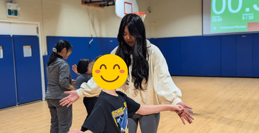 uby Bao, a volunteer with the SAAAC Autism Centre, engages with a child in a gymnasium. She is smiling warmly and extending her arms while guiding the child, who is wearing a black Sonic the Hedgehog t-shirt. Other participants and staff members are visible in the background, along with a basketball hoop and a digital timer on the wall.