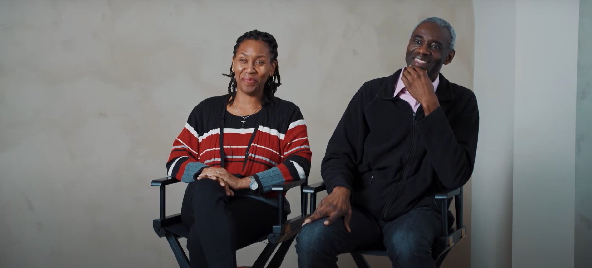 Sherron Grant and Clovis Grant, co-founders of the Sawubona Africentric Circle of Support, sit side by side in director's chairs, smiling warmly. Sherron wears a red, black, and white striped sweater, while Clovis wears a black jacket over a pink collared shirt. Both are parents of a son on the autism spectrum and have dedicated themselves to creating a support space for Black communities impacted by autism. The background is a neutral beige wall, and they appear engaged in conversation.