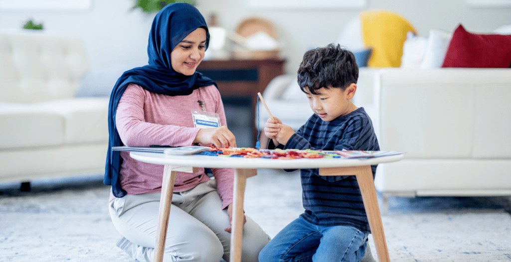 A therapist wearing a name badge and headscarf sits on the floor beside a young boy at a low table, engaging in a hands-on activity with colorful craft materials. The interaction reflects one-on-one ABA therapy in a warm, home-like setting, representing the supportive services provided at the SAAAC Autism Centre.