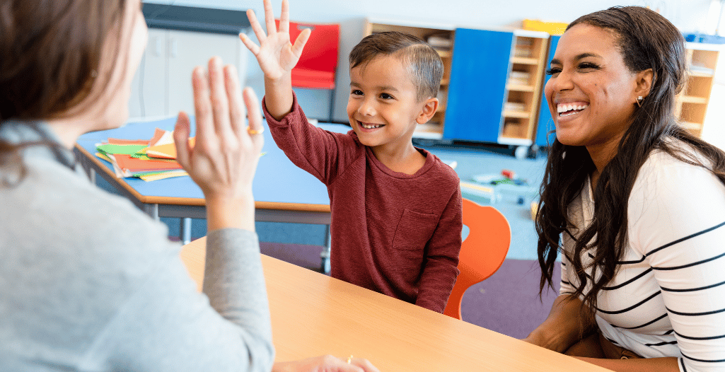 A smiling young boy sits between two adults at a table in a brightly lit classroom. The child enthusiastically raises his hand for a high-five with one adult, while the other adult, likely a caregiver, looks on with a warm smile. The scene captures positive interaction, learning, and supportive engagement—highlighting the collaborative and family-centered approach of the SAAAC ABA Suite’s Caregiver-Mediated Programs.