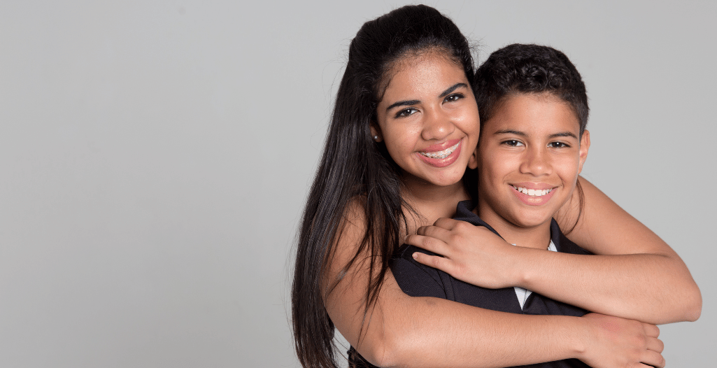 A smiling teenage girl with braces hugs her younger brother from behind, both looking happy and close. This image represents sibling relationships, family bonds, and support—reflecting the experiences of siblings of autistic individuals in immigrant families, the focus of the research study.