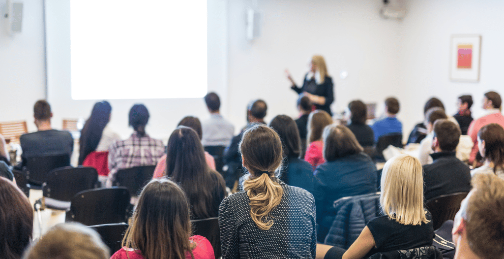 A diverse group of people attending a workshop or seminar in a conference room. The audience is seated, facing a presenter at the front of the room who is gesturing towards a projected screen. The speaker, a woman dressed in black, is slightly blurred in the background while the focus is on the backs of the attendees.