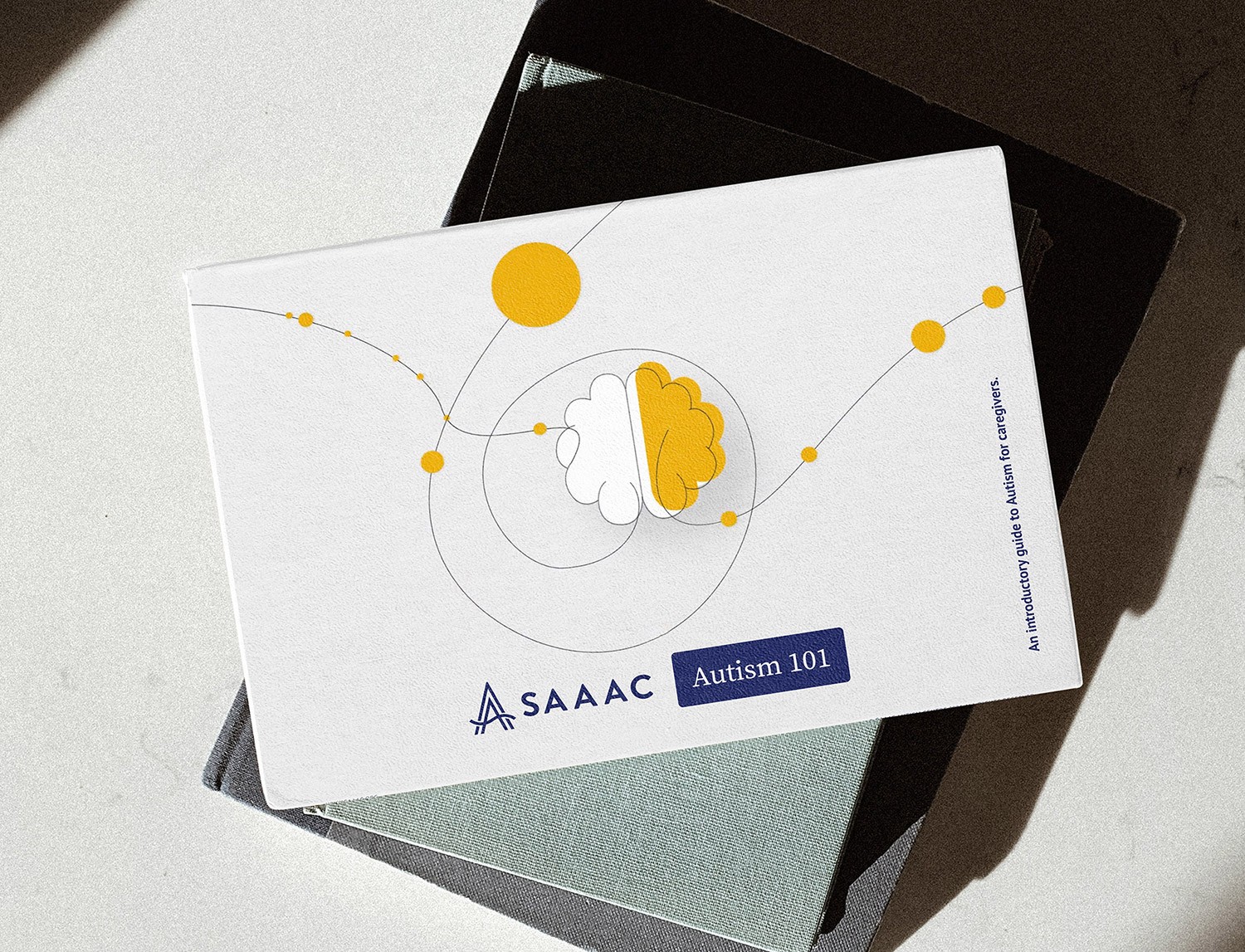 A booklet titled 'Autism 101' by the SAAAC Autism Centre rests on a textured surface, partially inside a dark folder. The cover features a minimalist illustration of a brain with yellow and white hemispheres, surrounded by orbit-like lines and yellow circles. The subtitle reads, 'An introductory guide to Autism for caregivers.