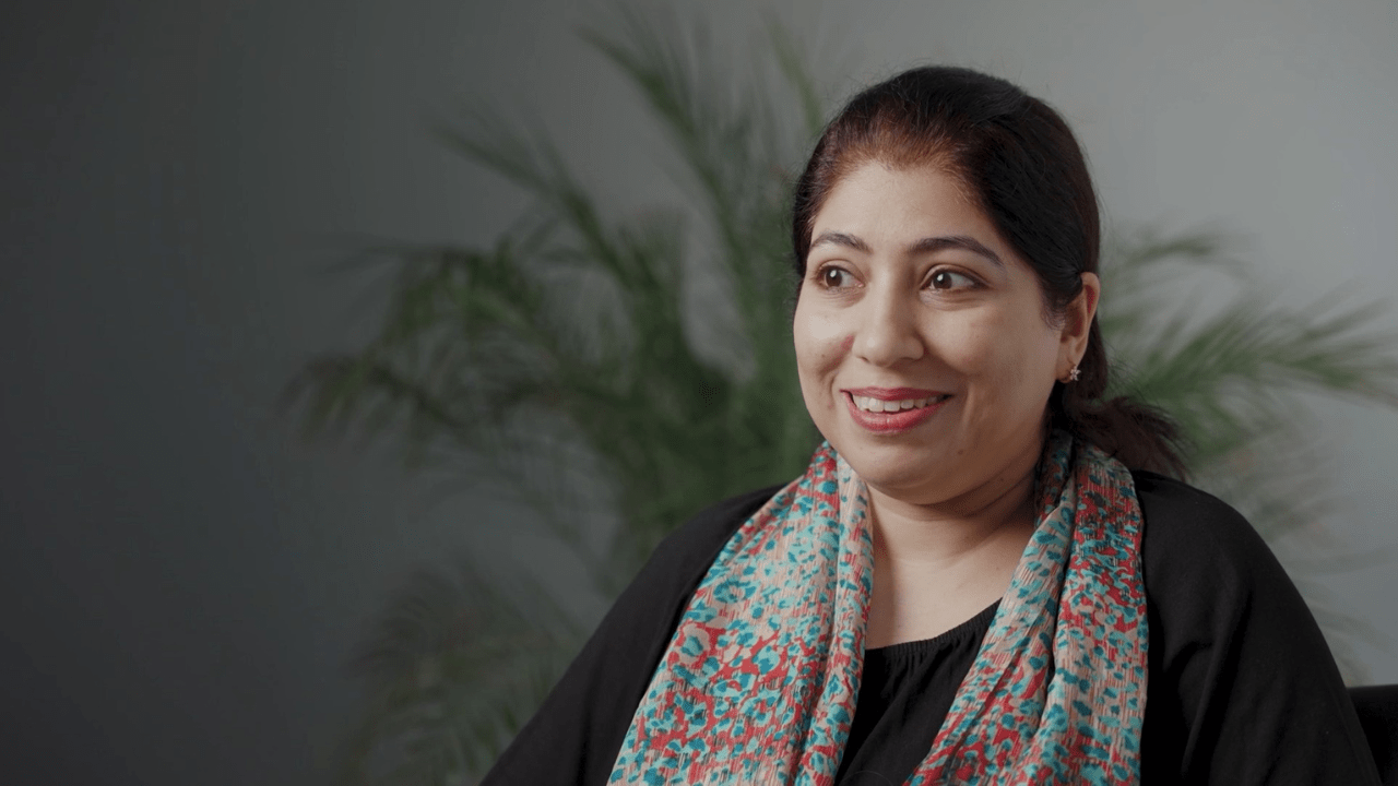 Quratulain, smiling warmly while seated in a softly lit room with a plant in the background, participates in SAAAC Autism Centre's Autism 101 campaign to support awareness and acceptance of autism.