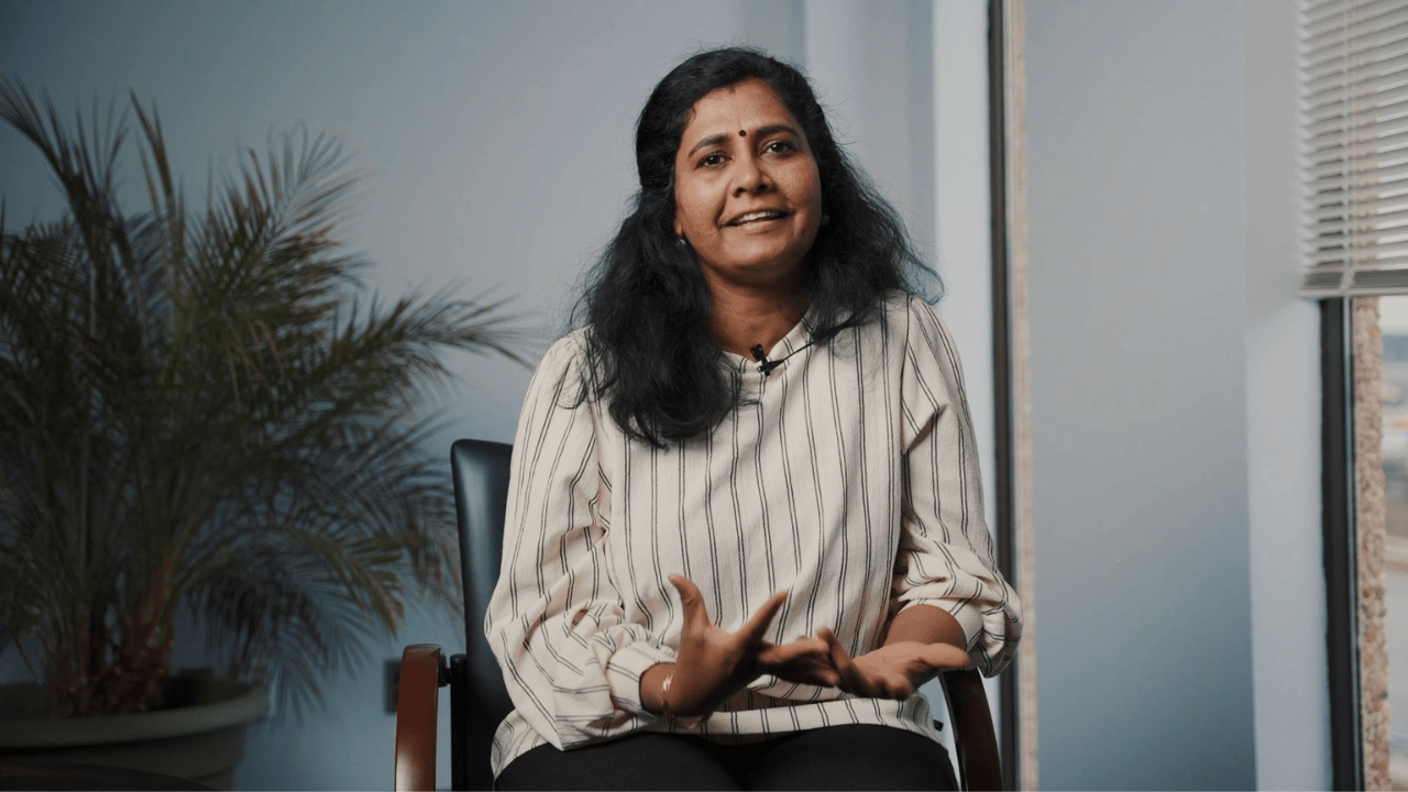 Yalini Murugatheeswaran, a woman with long black hair, sits in a chair wearing a striped blouse, sharing her personal journey of raising a child diagnosed with autism. She has a thoughtful expression as she speaks, with a calm office setting in the background, including a potted plant and a window with blinds partially open.