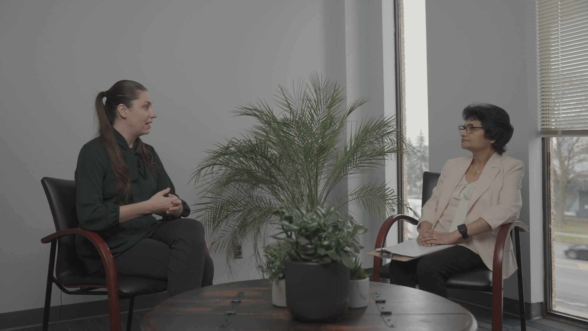 amie Laframboise and Lakshmi Solomons are seated in a professional office setting, engaged in a conversation about autism. Jamie, wearing a dark green blouse, gestures with her hands as she speaks, while Lakshmi, dressed in a light pink blazer, listens attentively with a clipboard in her lap. A round wooden table with potted plants is in the foreground, and large windows with blinds partially open reveal an outside view of a street with trees and buildings. The room has a neutral-toned wall and a calm, professional atmosphere.