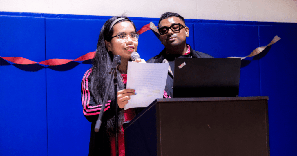 An artist on the autism spectrum speaks at a podium, holding a microphone and a printed speech, during an event at the SAAAC Autism Centre. She stands in front of a laptop with a supportive individual beside her. The background features a blue padded wall decorated with a red and beige twisted ribbon garland.