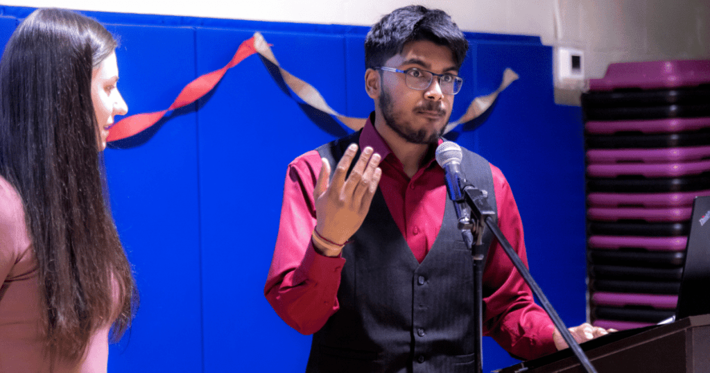 Vignesh Athavan emceeing the Autism Acceptance Day celebration at the SAAAC Autism Centre, speaking at a podium with a microphone while gesturing with one hand. A woman stands beside him, and festive streamers decorate the background.
