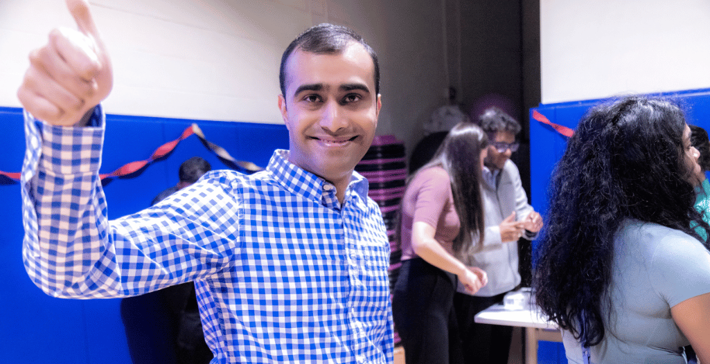 Kirishan Thananjeyan, an autistic musician and instrumentalist, smiles and gives a thumbs-up while attending the SAAAC Autism Centre's Autism Acceptance Day event. He is wearing a blue and white checkered shirt, with people engaging in activities in the background against a festive setting.
