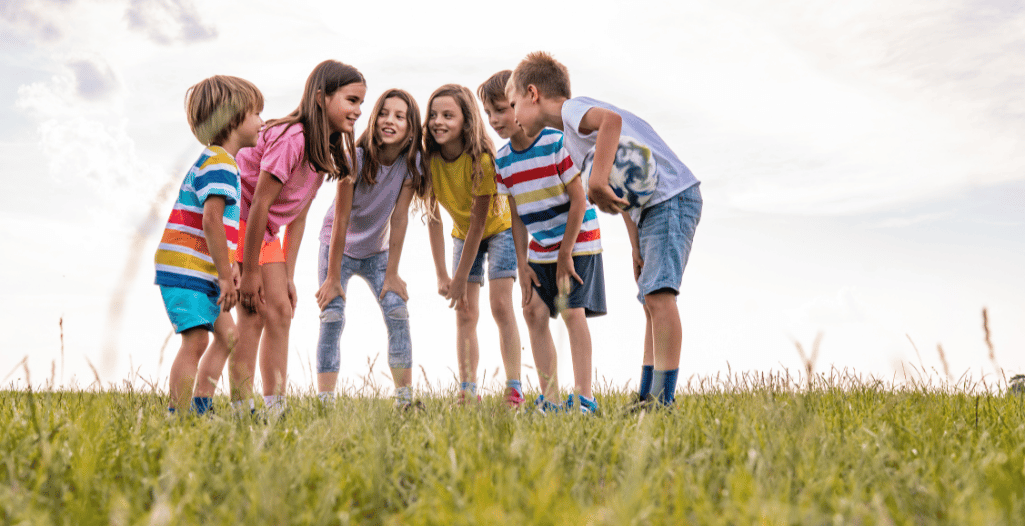 A group of six children standing in a circle on a grassy field, smiling and chatting with each other. They are dressed in colorful casual summer clothes, and the sky is bright and slightly cloudy in the background. The mood is cheerful and playful, suggesting outdoor social interaction.