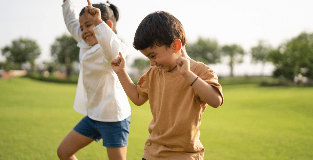Two young children smiling and playing energetically on a grassy field, with trees in the background. The boy wears a light brown t-shirt and the girl wears a white shirt and denim shorts. Both have their arms raised in joyful motion, capturing a moment of outdoor fun and movement.