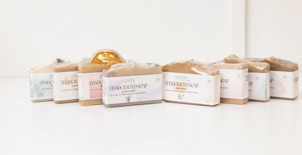 A collection of Mindful Glow handcrafted soaps arranged in a diagonal line on a white surface. Each soap bar features unique natural ingredients and is wrapped in minimalist labels indicating scents like Calming Lavender and Lemon Turmeric.